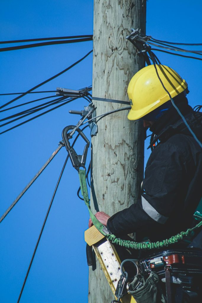 man-in-black-jacket-and-yellow-hard-hat-climbing-on-brown-wooden-post-during-daytime-jhgfclznouy